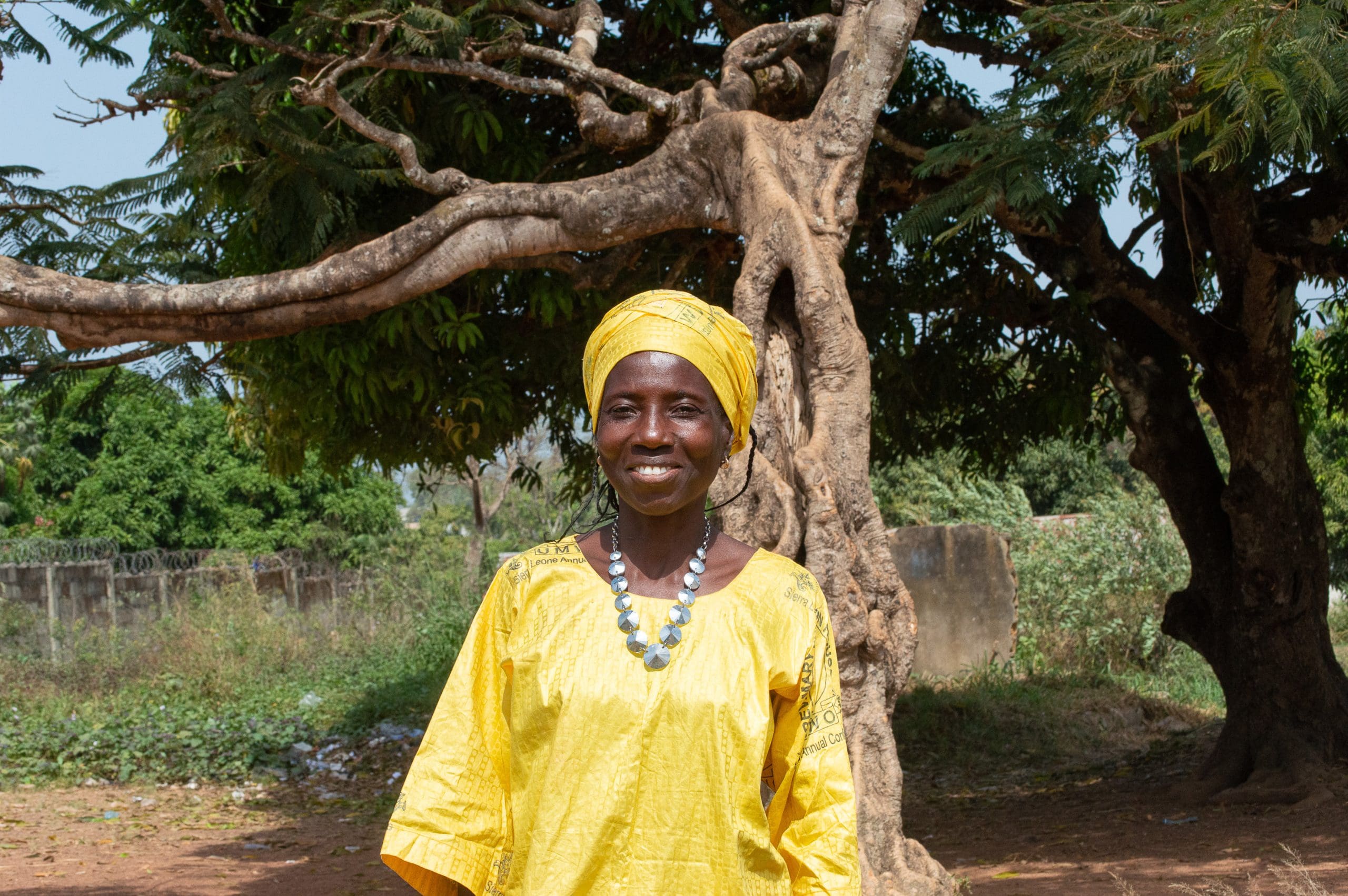 A smiling woman in a yellow outfit and head wrap standing under a tree in Sierra Leone